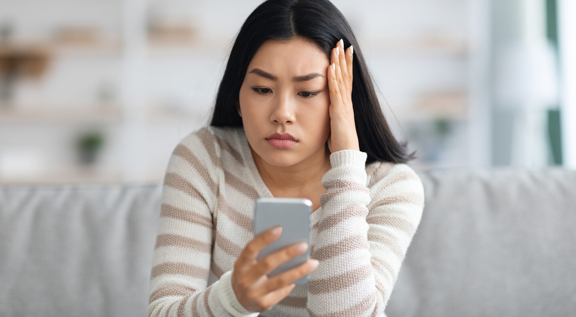 Bad News. Upset Asian Female Holding Smartphone, Looking At Mobile Phone Screen With Worry While Sitting On Couch At Home, Anxious Korean Woman Touching Head While Reading Unplesant Message, Closeup