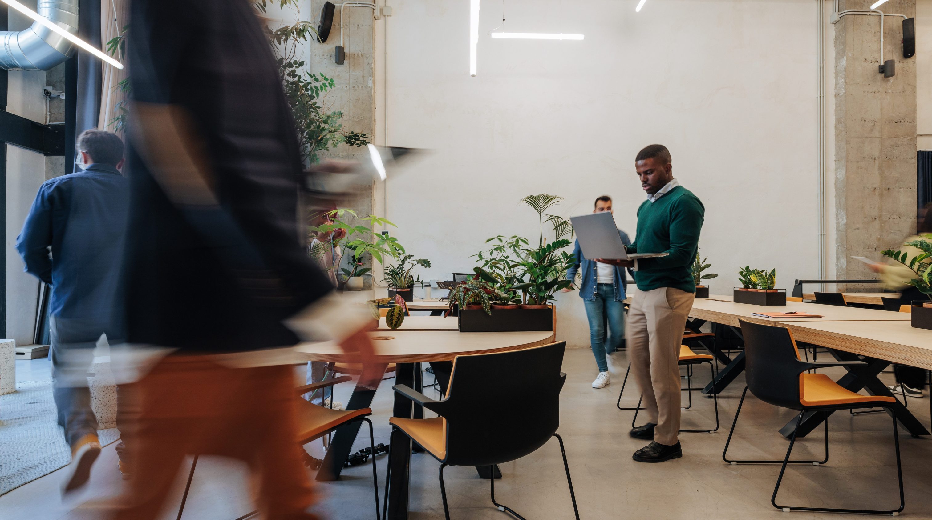 A spacious Madrid coworking area with plants and long desks. An African American man stands with a laptop while colleagues move in blur, capturing dynamic, collaborative office atmosphere.